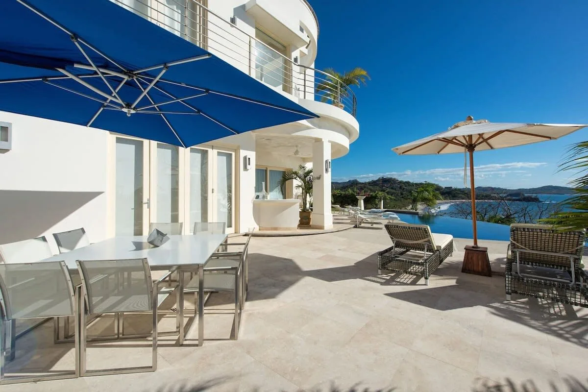 A sunlit outdoor terrace has a dining table, lounge seating, and views of the water.