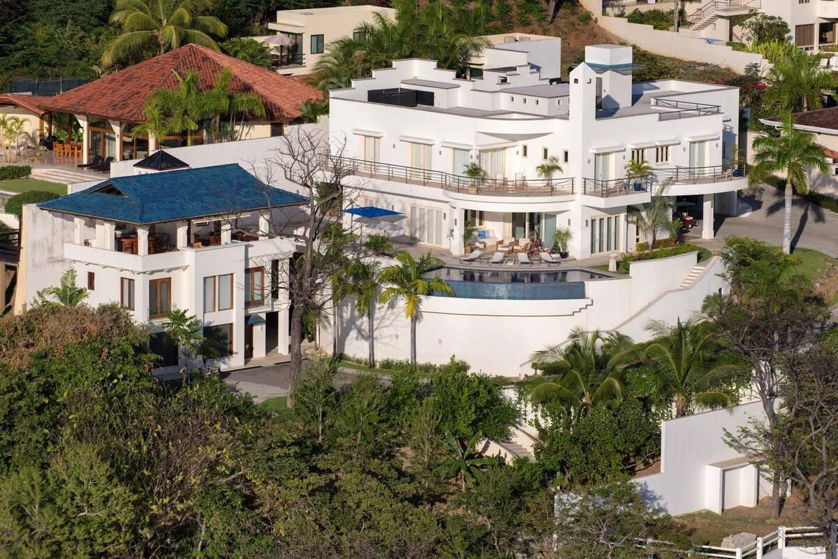 A modern white residence with balconies and a curved pool, surrounded by greenery.