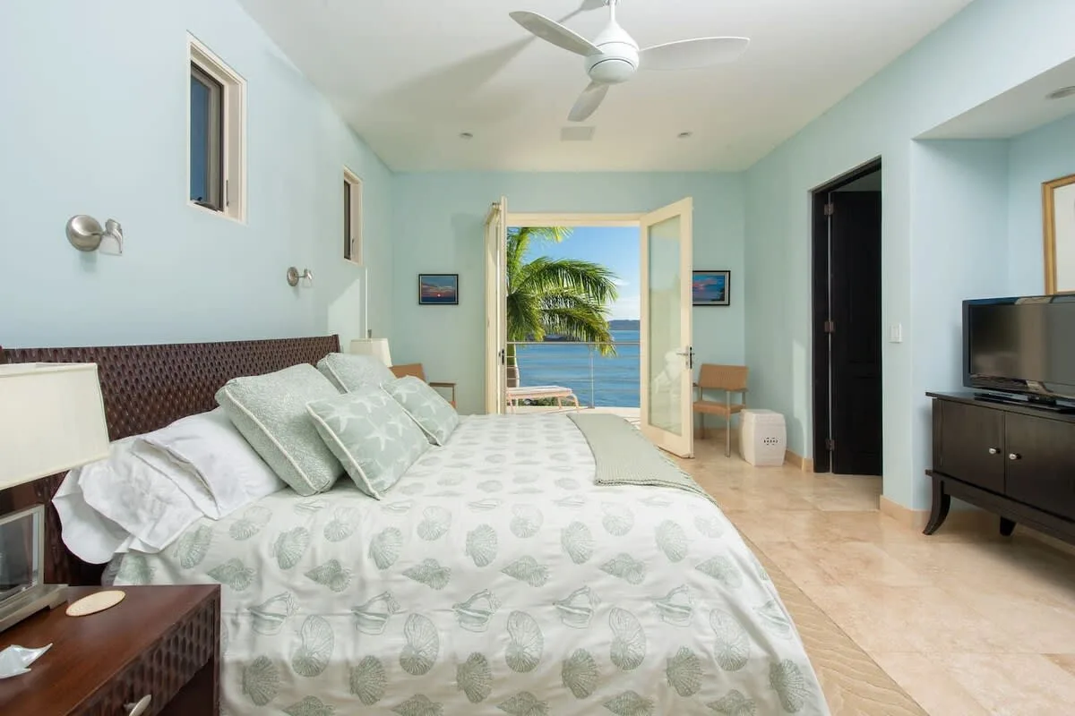 A bright bedroom with a large bed and open doors facing palm trees and water.