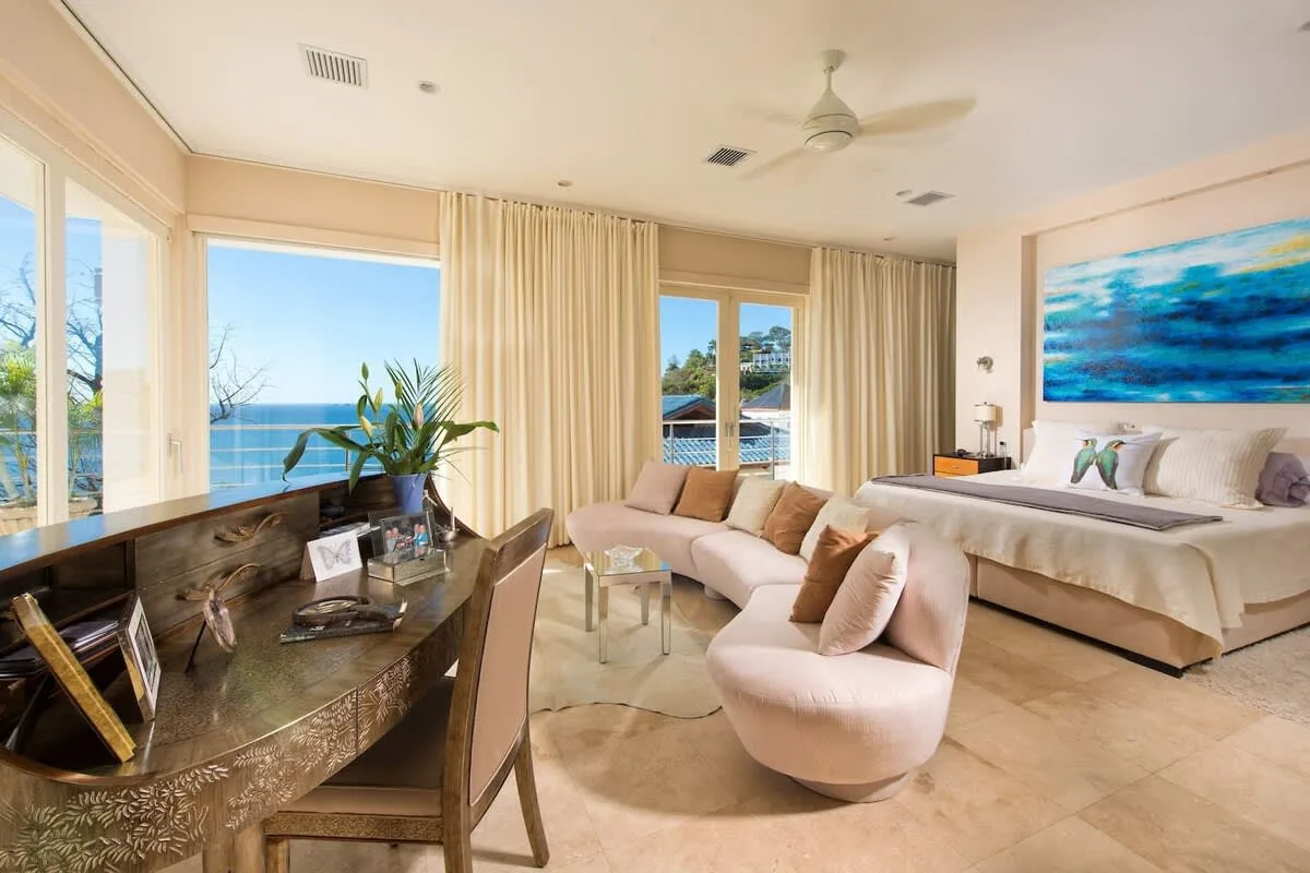 A bright bedroom with a bed, seating area, and large windows overlooking the water.