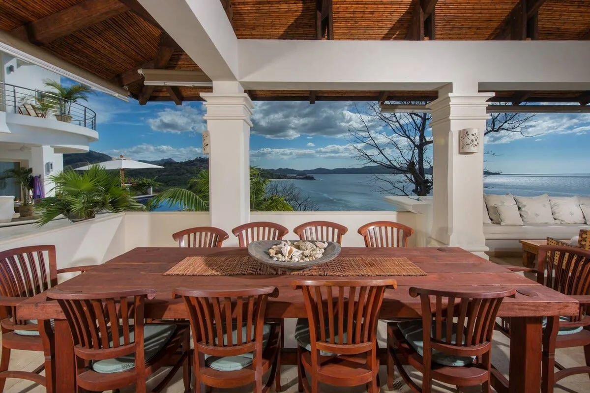 An outdoor dining area overlooks the water, with a long wooden table and chairs.