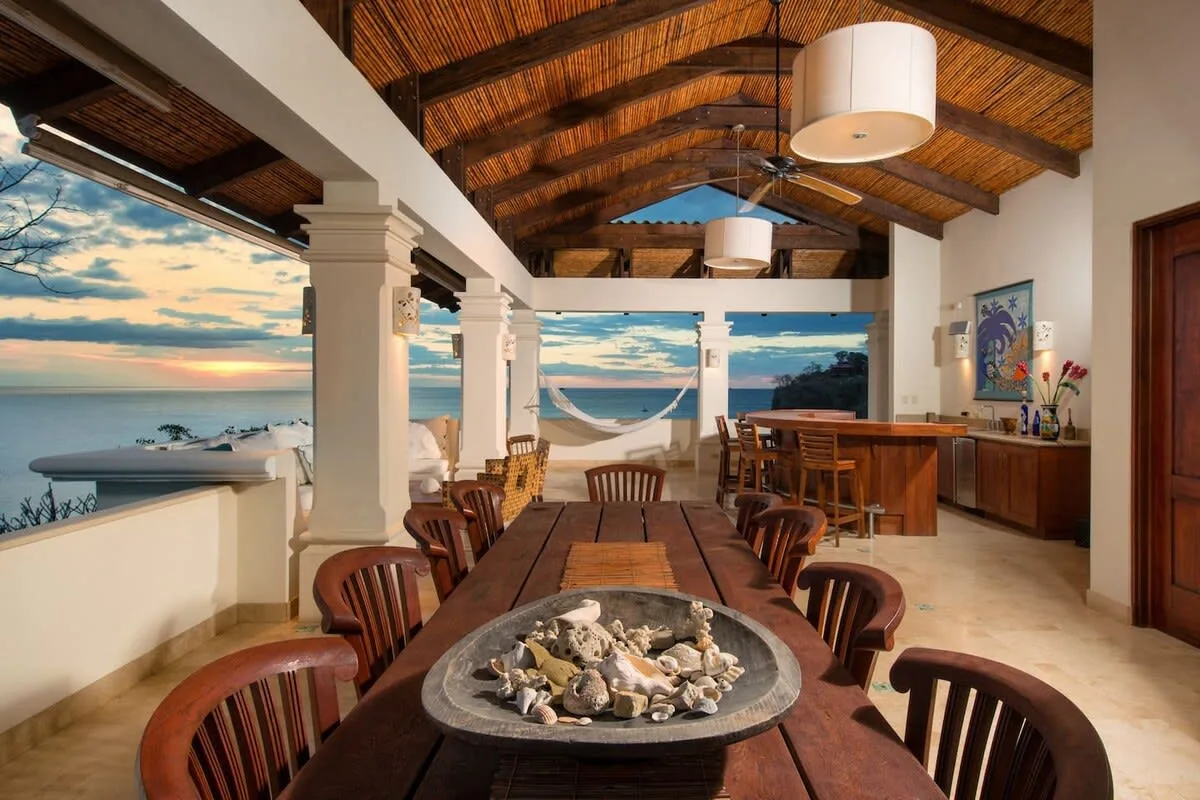 An open dining area with a long wooden table overlooks the ocean.