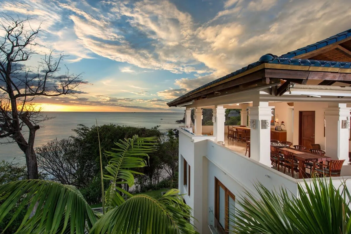 A covered dining terrace overlooks the ocean beneath a cloud-filled sunset sky.