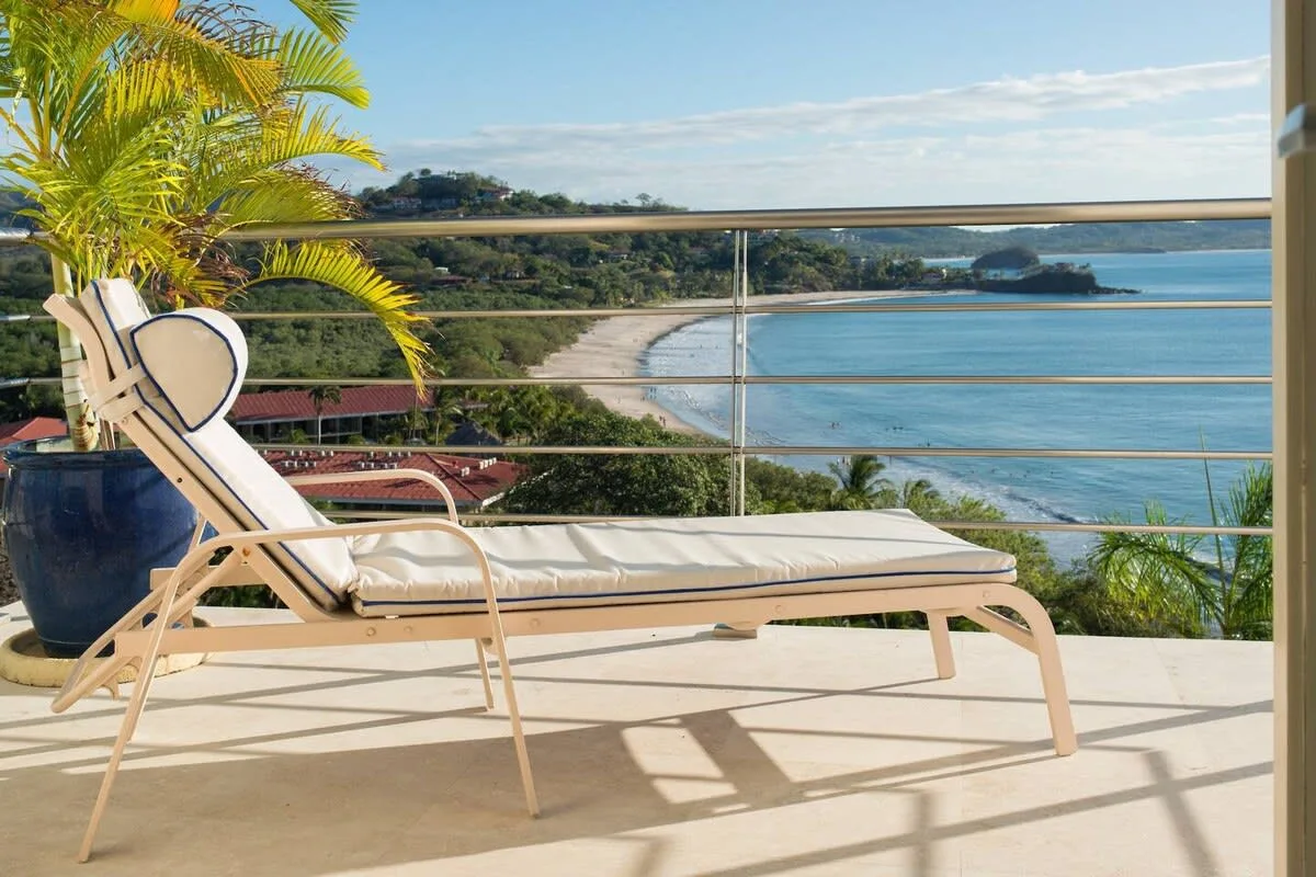 A sun lounger sits on a terrace overlooking a curved beach and blue water.