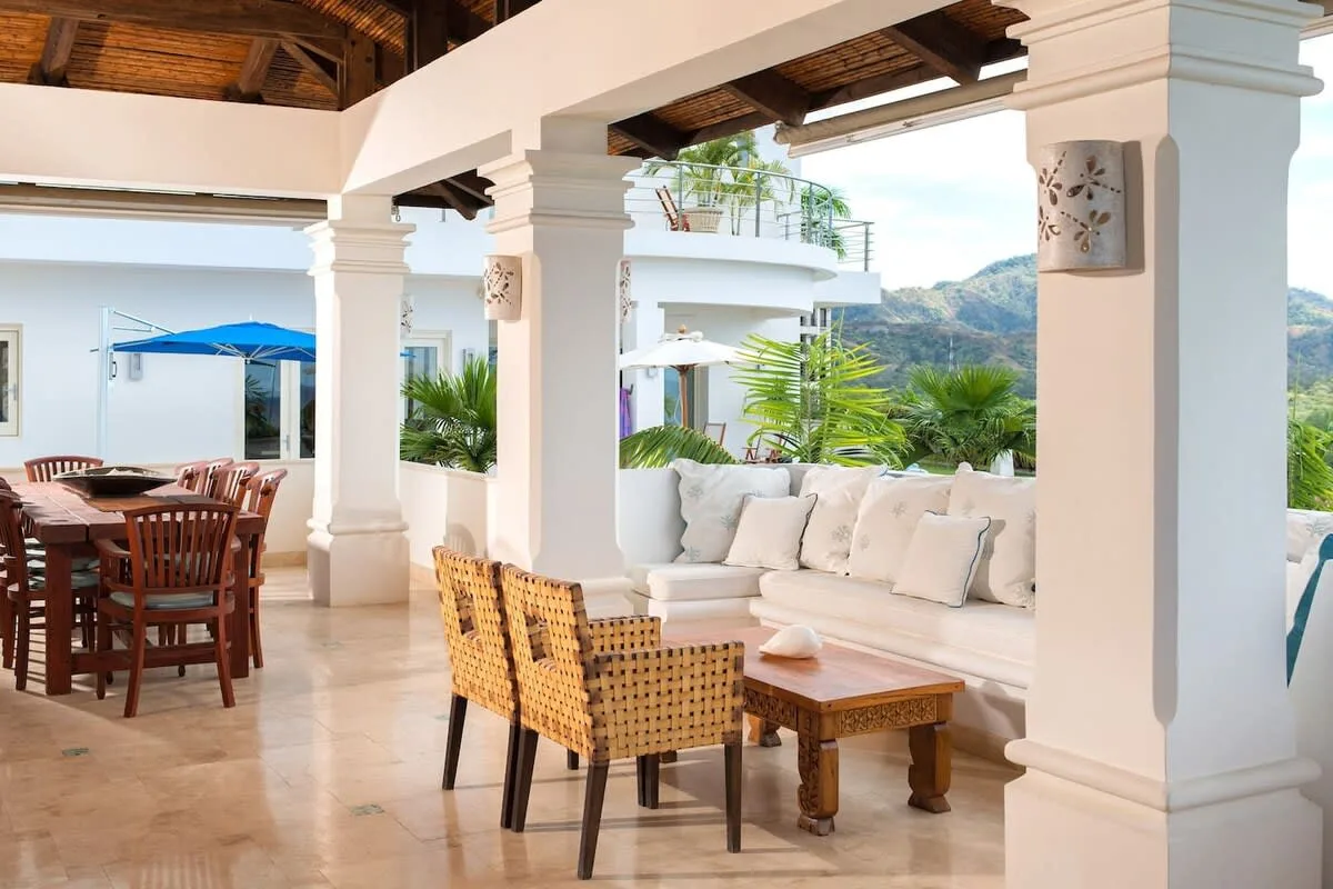 A covered patio with dining and lounge seating overlooks a green hillside.