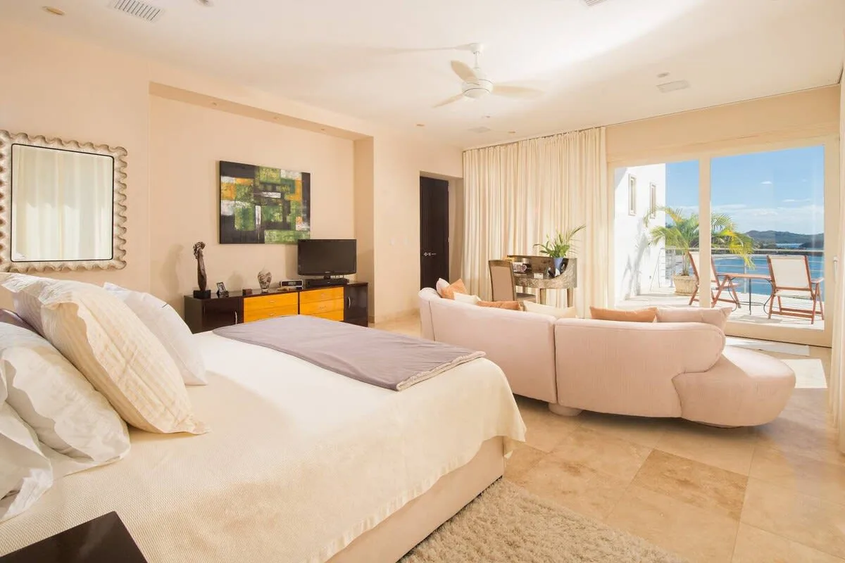 A bright bedroom opens into a sitting area beside large windows overlooking the water.