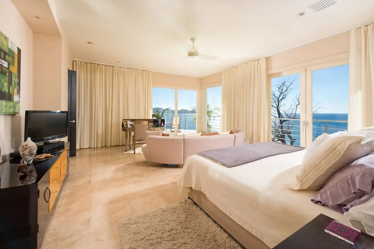 A bright bedroom opens onto a living area with large windows overlooking the ocean.