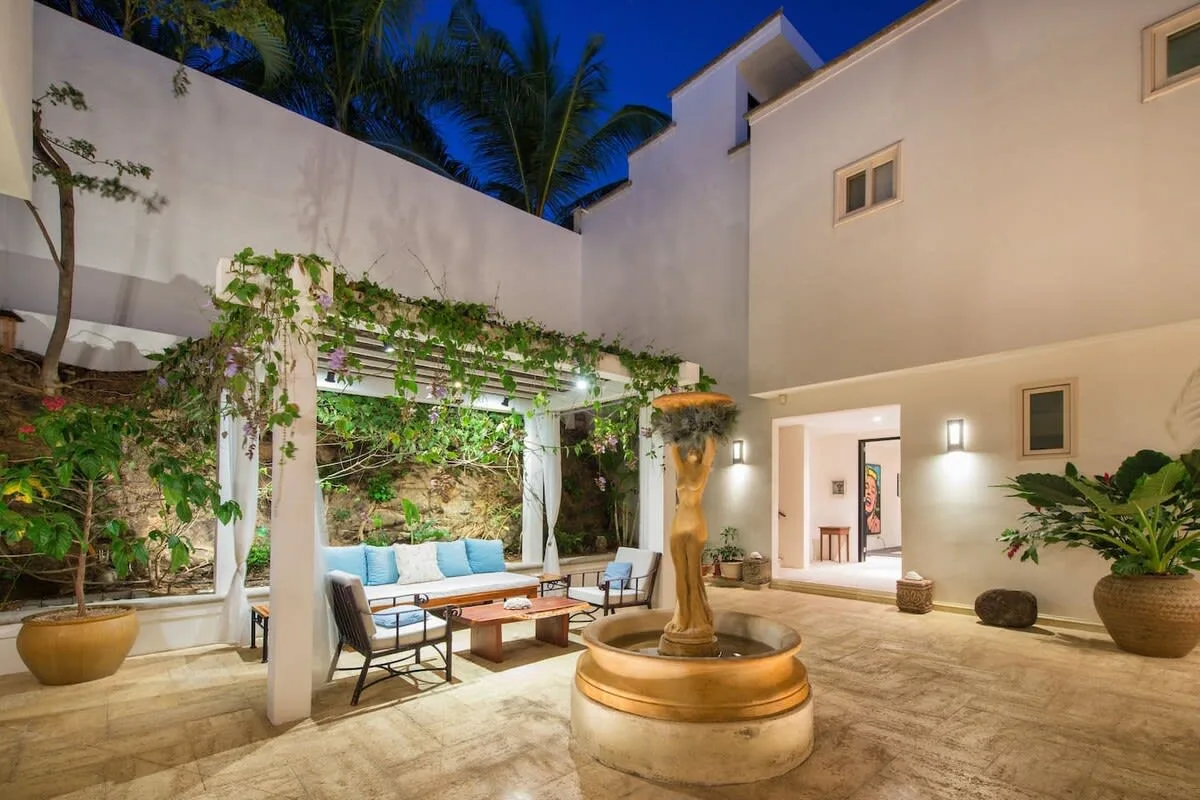 An outdoor courtyard features a central fountain and seating beneath a leafy pergola.