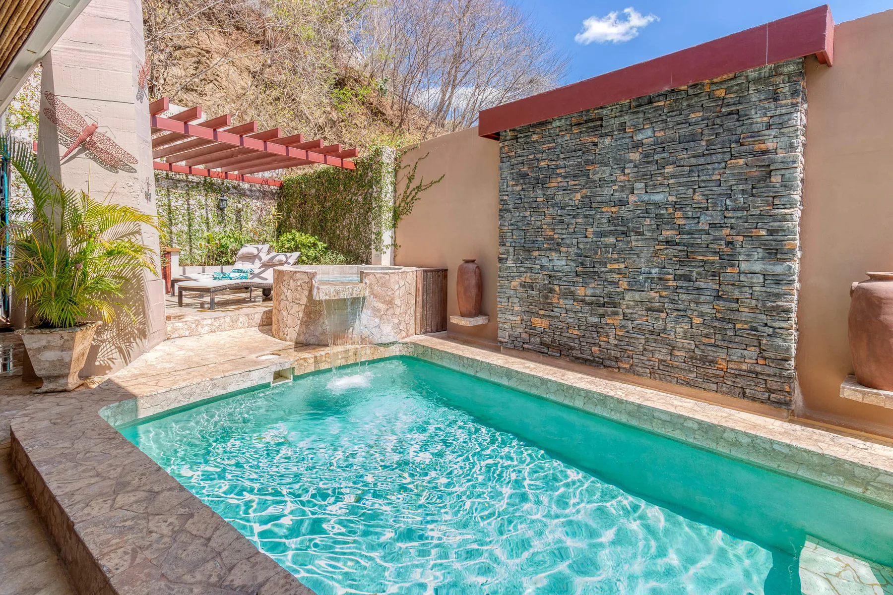 A small outdoor pool sits beside a textured wall and a shaded seating area.