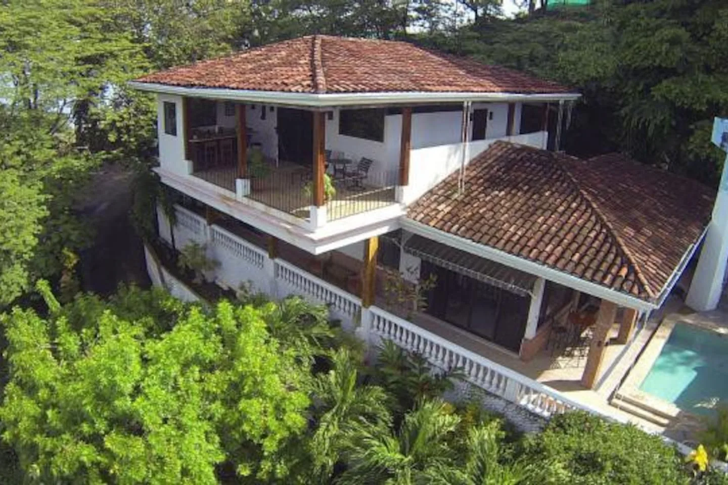 A two-story house with tiled roofs and a wraparound balcony stands among trees.