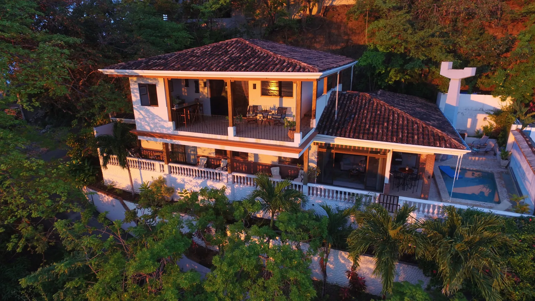 A two-story house with covered balconies sits among dense trees and gardens.