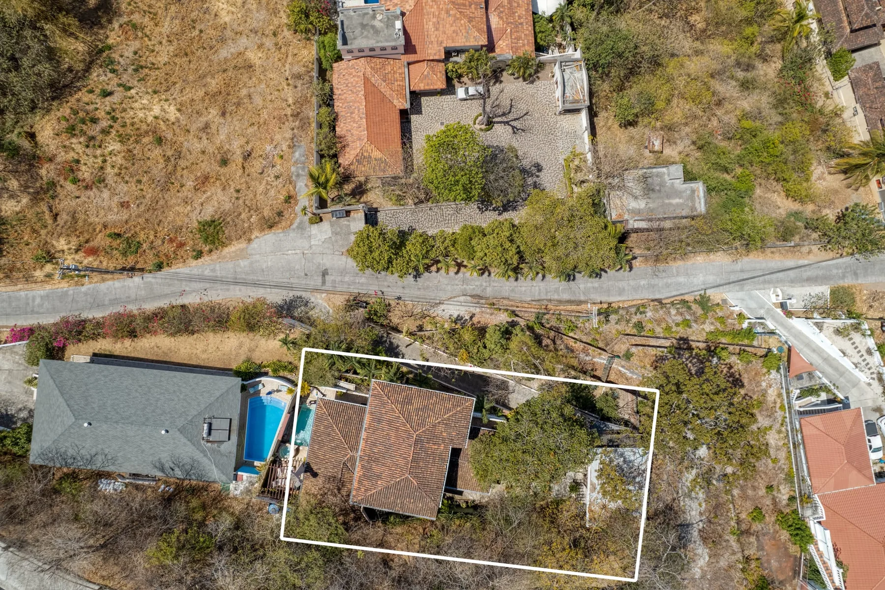 An aerial view shows a house and surrounding dry, scrubby ground beside a road.