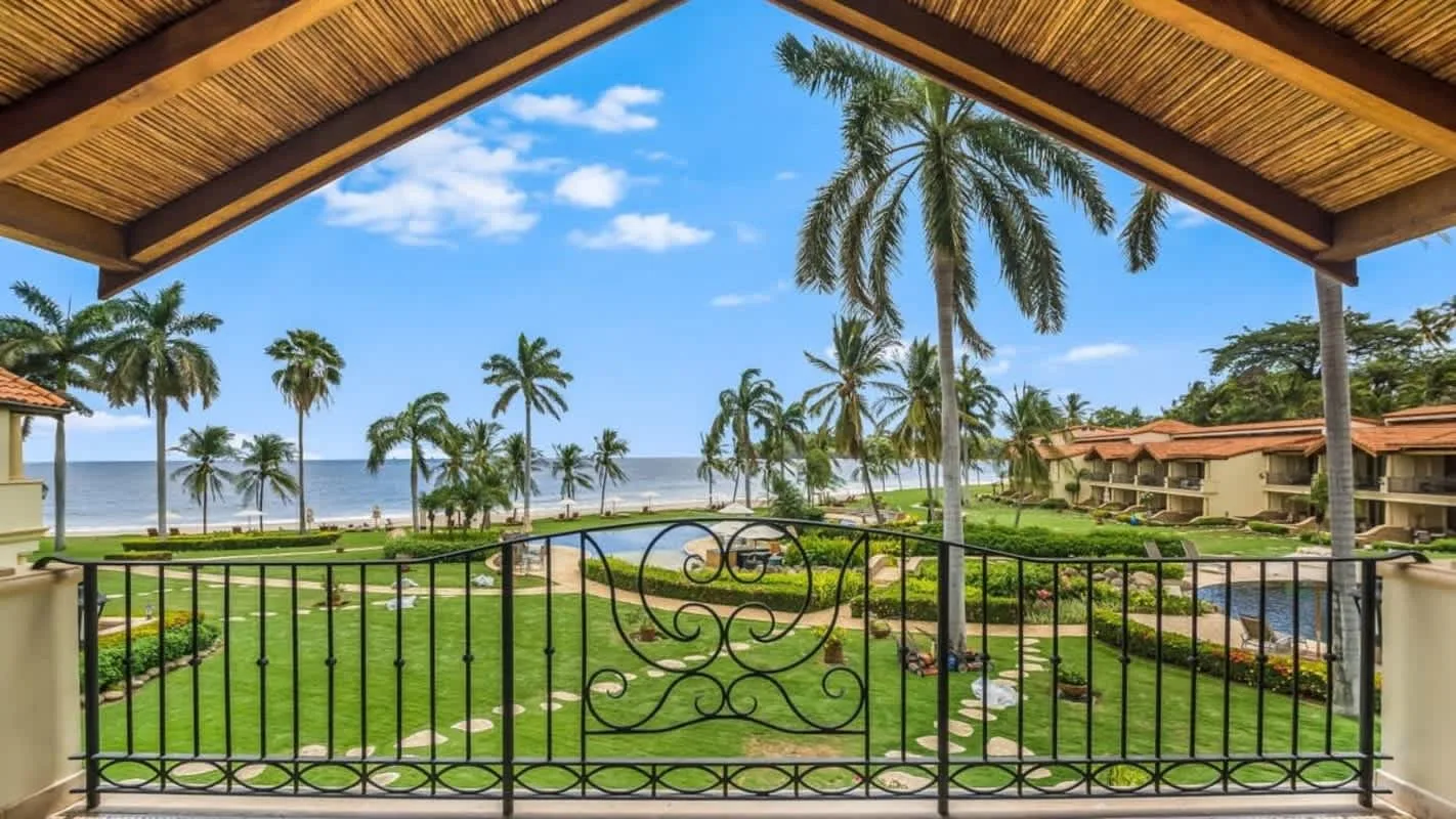 A covered balcony overlooks landscaped gardens, palm trees, and the ocean.