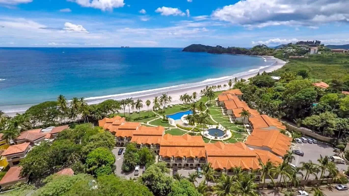 Shared amenity: Aerial view of a coastal resort with orange-roofed buildings beside a curved beach.