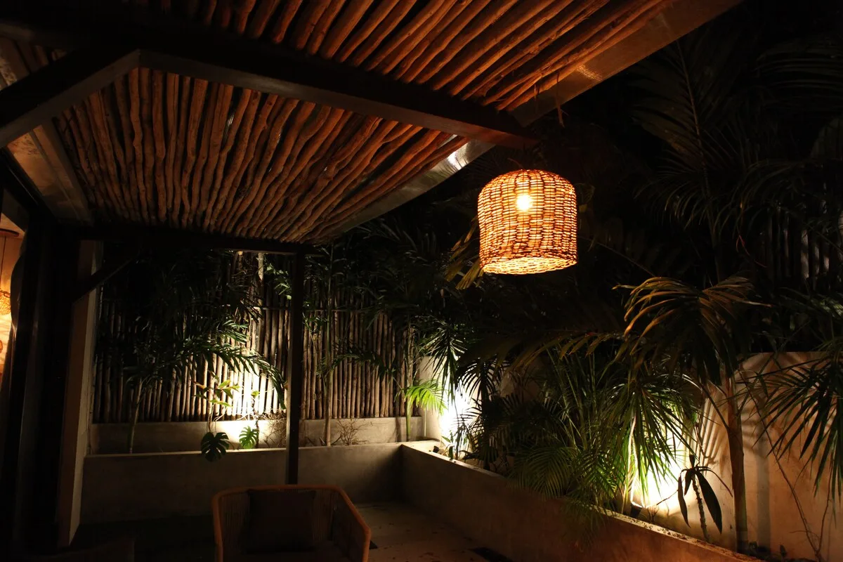 Un patio cubierto con poca luz, plantas tropicales y una lámpara colgante tejida.