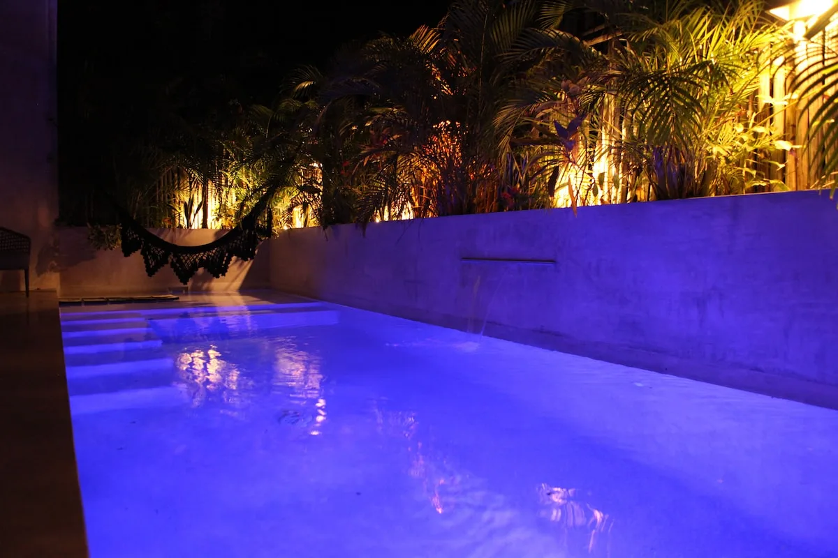 Una piscina iluminada de azul está junto a un muro bajo y plantas tropicales por la noche.