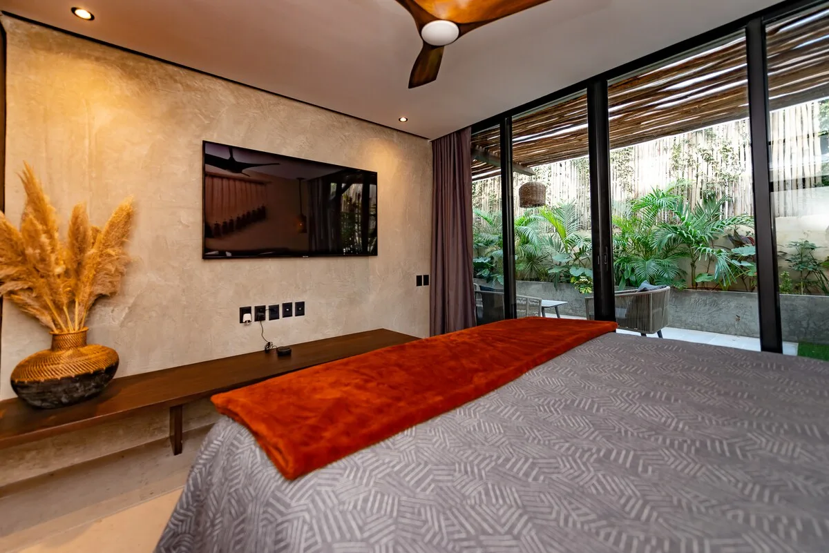 Un dormitorio con cama, televisor en la pared y puertas de vidrio frente a la vegetación.