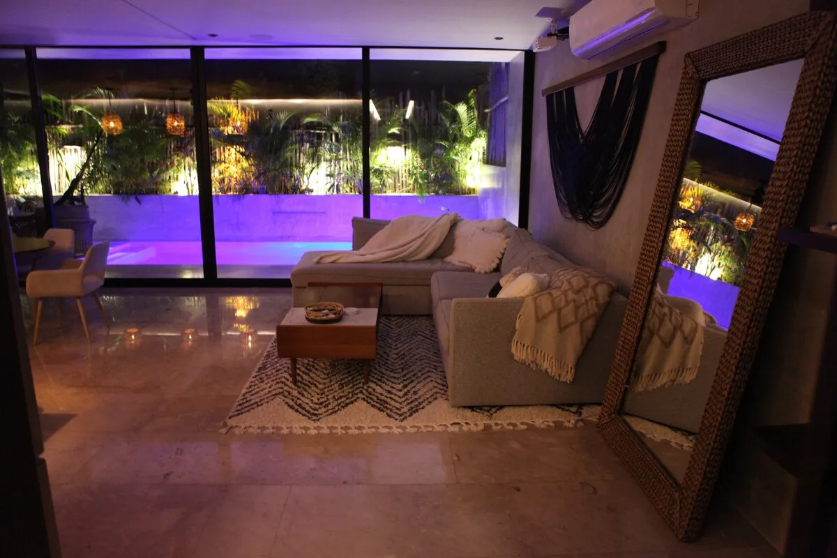 Una sala de estar con asientos y una mesa baja mira hacia grandes ventanales con vegetación iluminada.