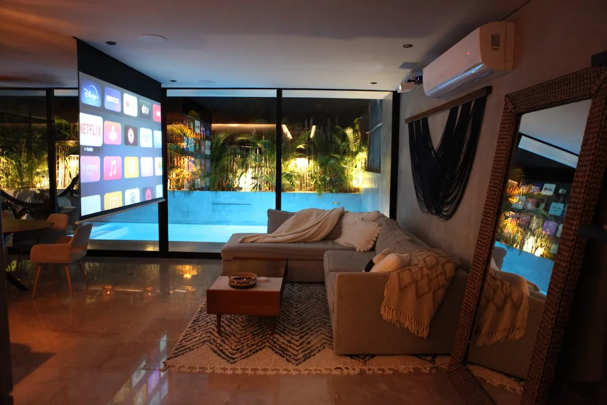 Una sala de estar con asientos seccionales, una pantalla grande y ventanas con vista a la vegetación.