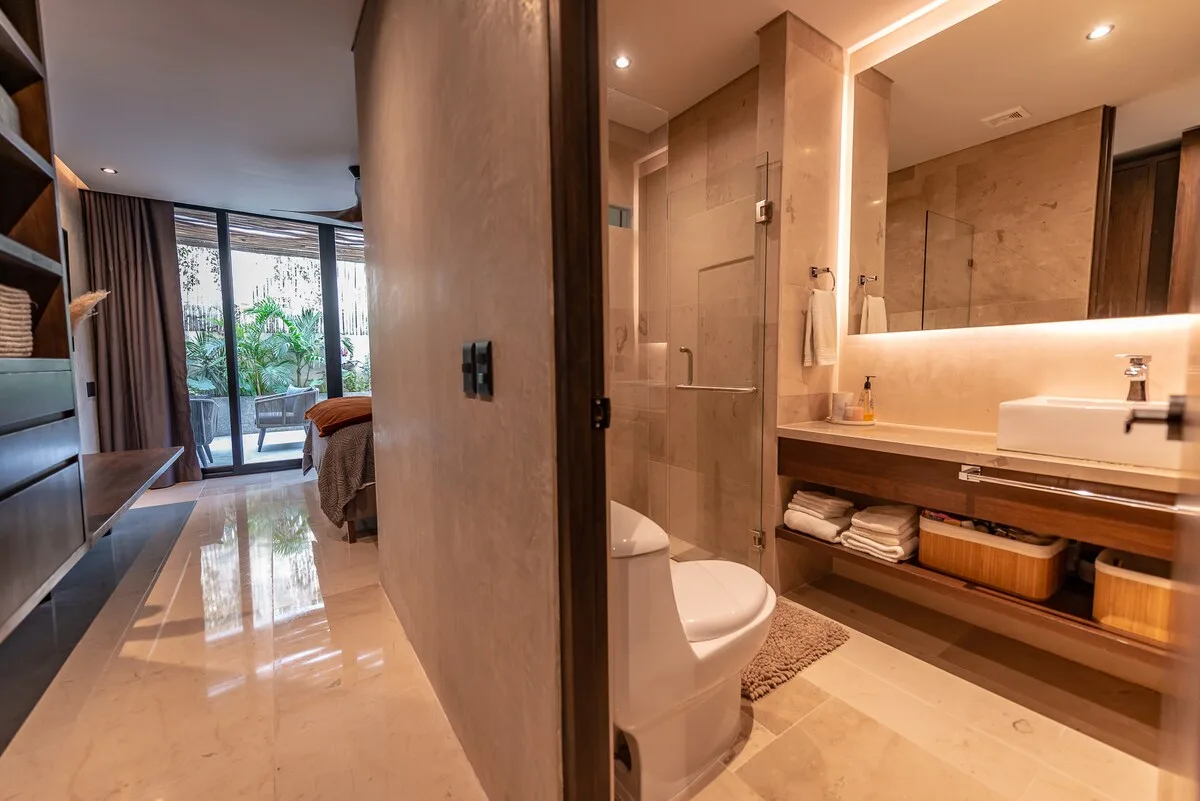 Un baño con lavabo e inodoro se abre a un dormitorio con ventanales.