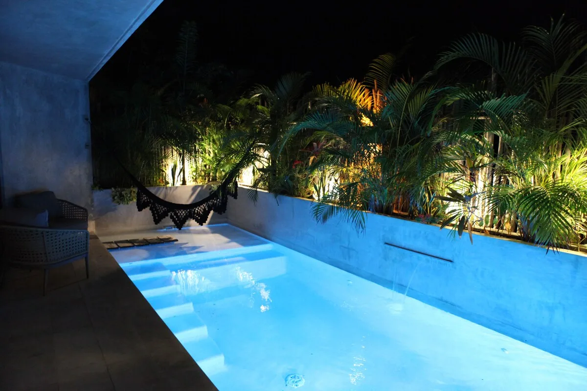 Una piscina iluminada por la noche junto a plantas frondosas y una hamaca.