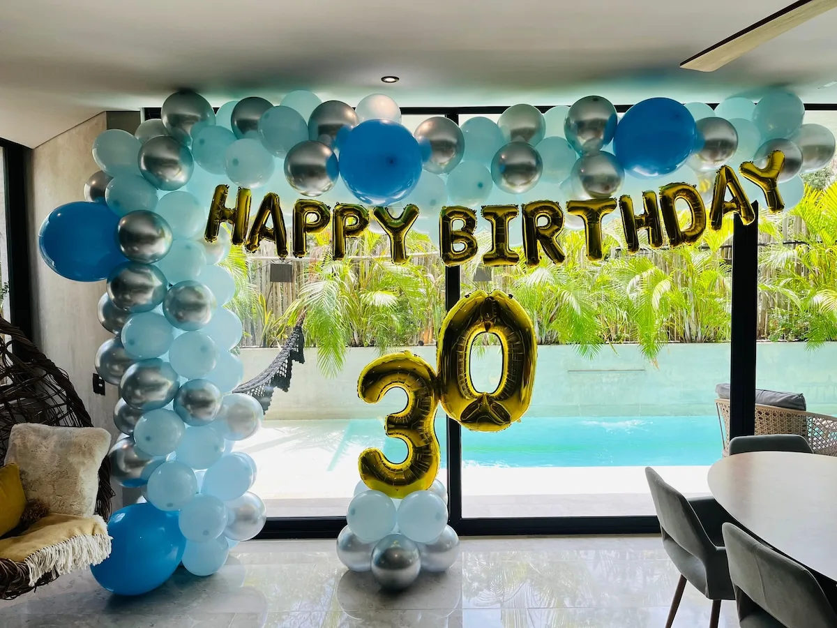 Una decoración de cumpleaños con globos enmarca una sala luminosa frente a una piscina.