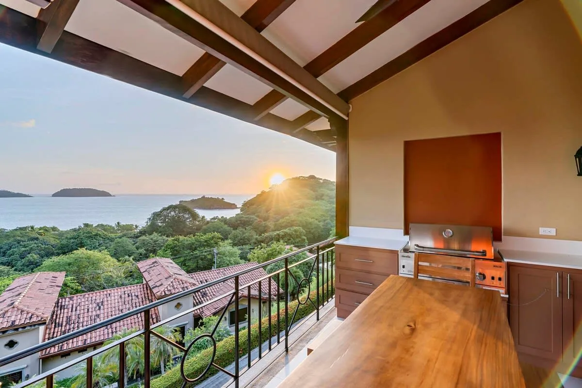 A covered balcony overlooks coastal rooftops and the ocean at sunset.