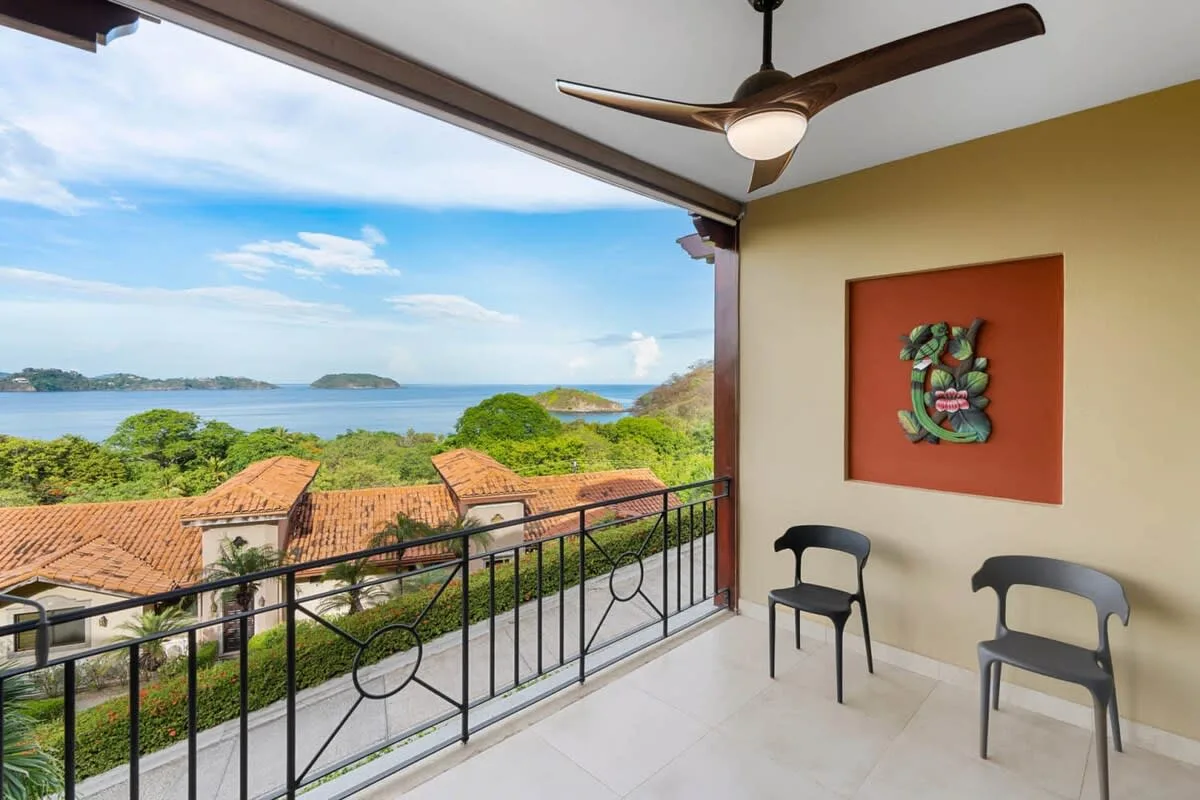 A covered balcony overlooks greenery, terracotta rooftops, and the sea.
