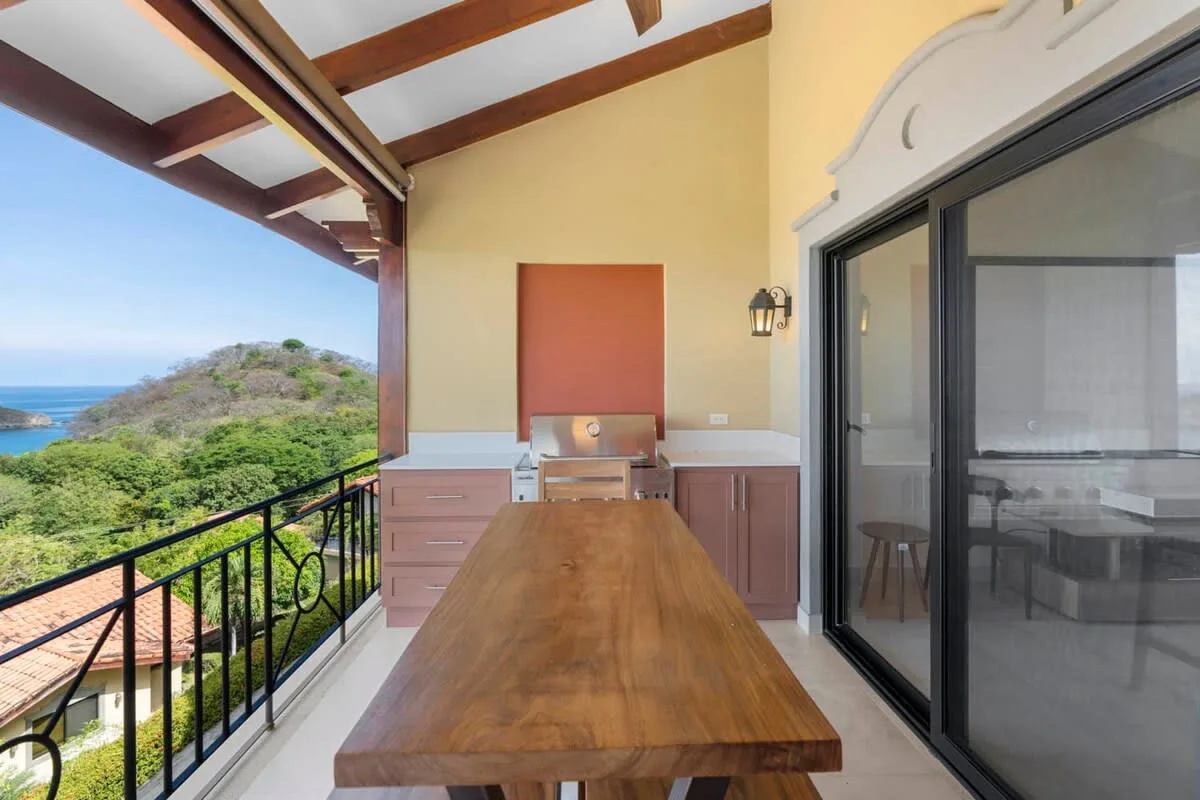 A covered balcony dining area with a wooden table overlooking the ocean and hillside.