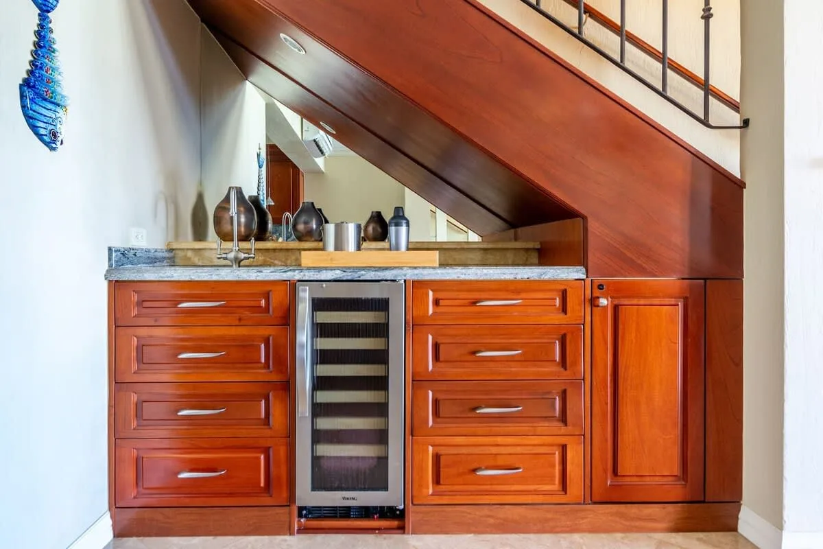 A built-in bar with wooden cabinets, a countertop, and a wine cooler beneath the stairs.