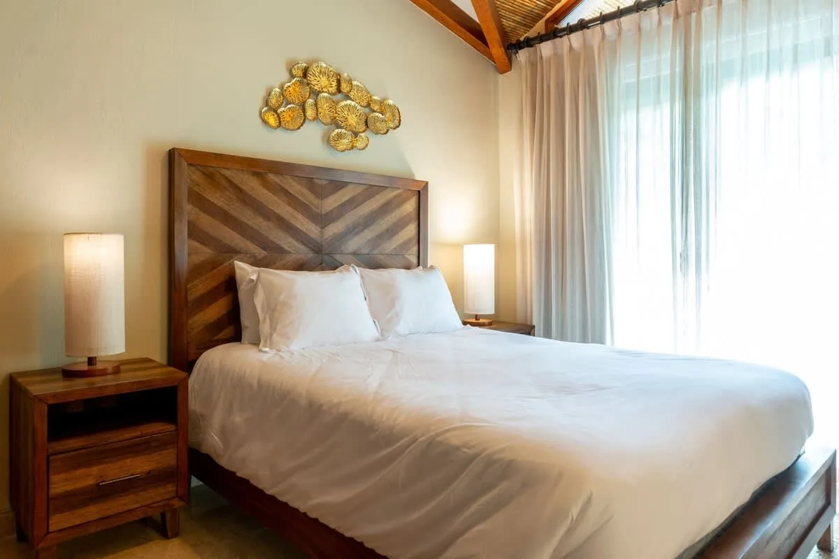 A bedroom with a large bed, wooden headboard, and bright curtained window.