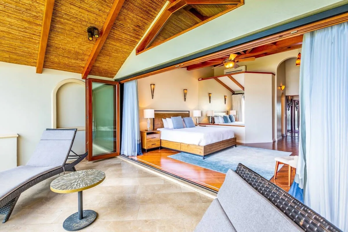 A bedroom with a king-sized bed opens onto a sitting area beneath a wooden vaulted ceiling.