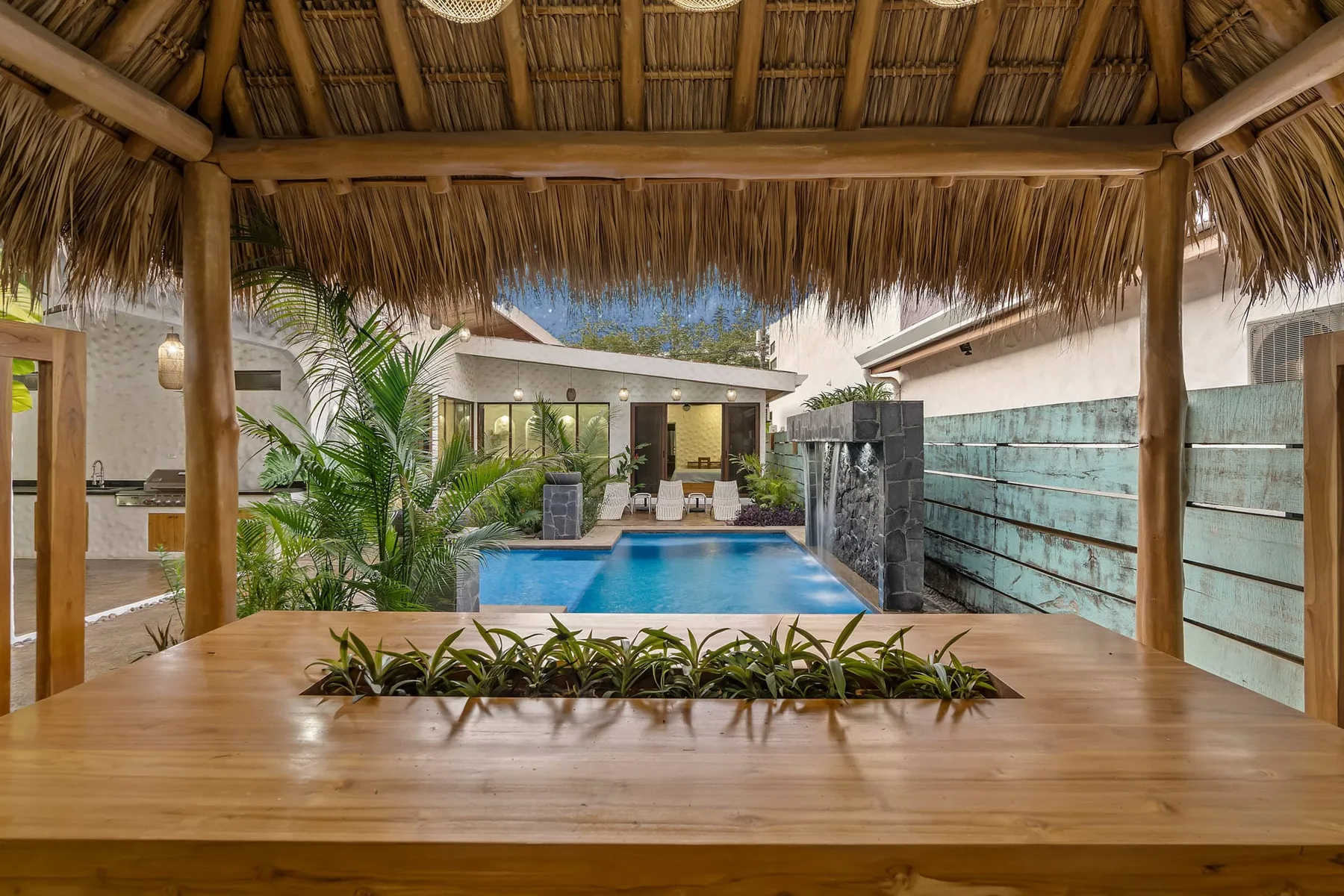 Zona de piscina al aire libre bajo un techo de palma, con plantas tropicales y asientos de madera.