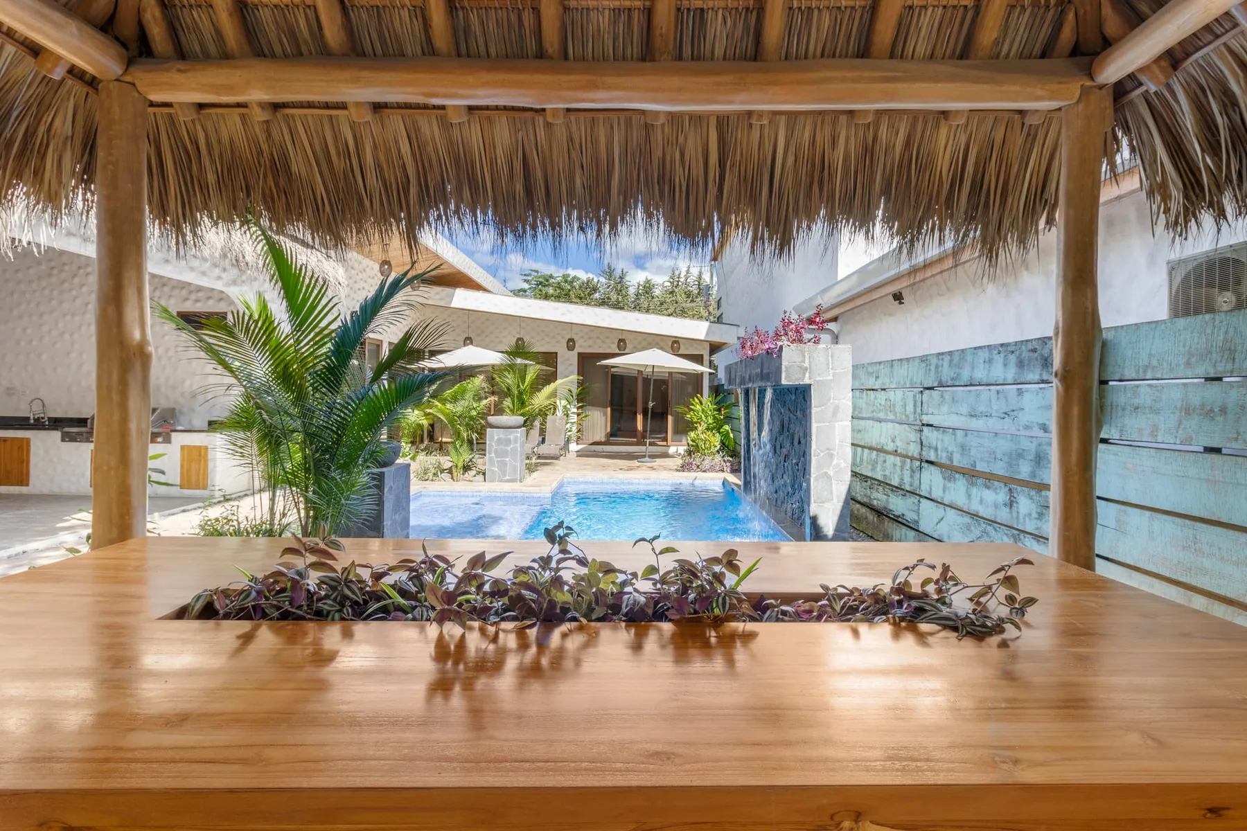 Una piscina y plantas tropicales se ven más allá de una terraza de madera sombreada.