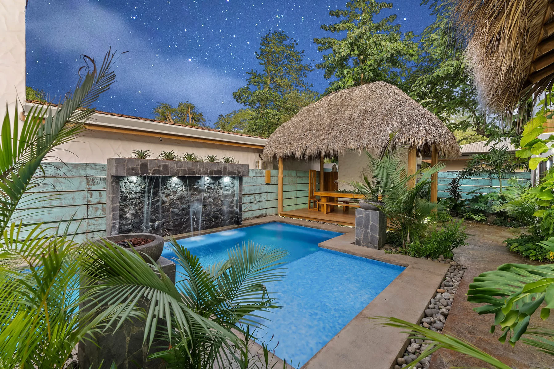A tropical pool courtyard with a thatched-roof shelter and lush plants.
