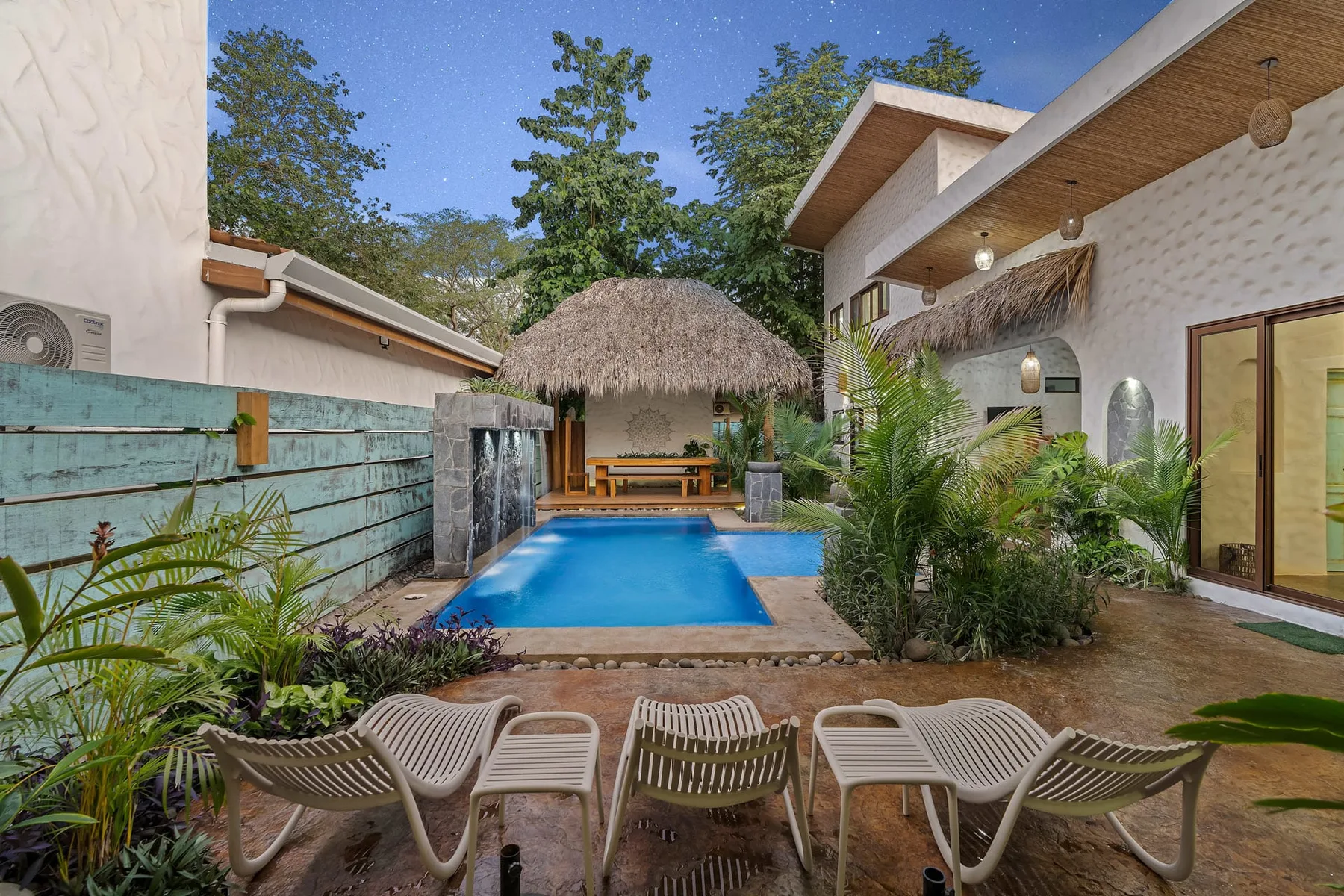 Una piscina exterior está junto a una estructura de sombra con techo de palma y una zona ajardinada con asientos.