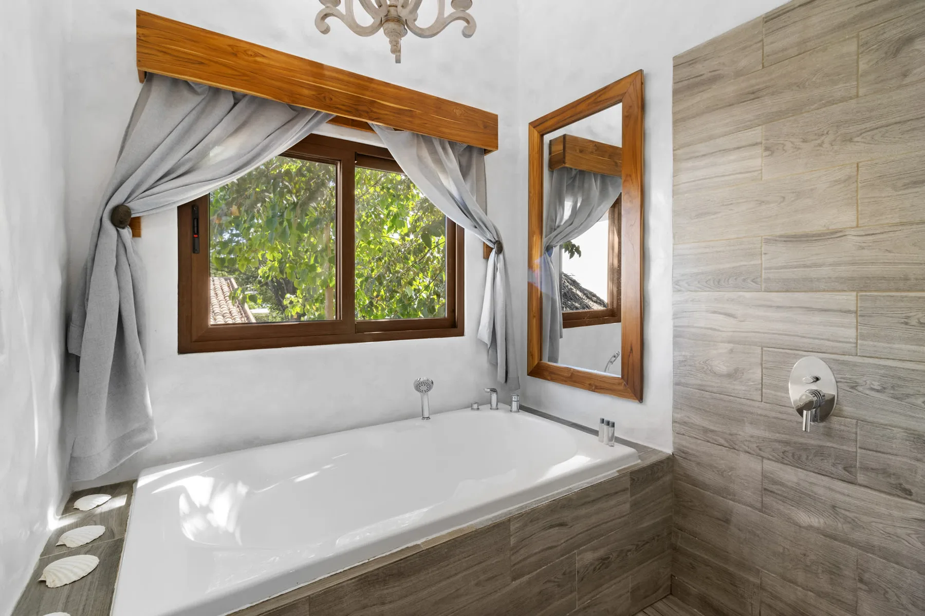 Un baño con bañera bajo una ventana con cortinas y una zona de ducha revestida de azulejos.