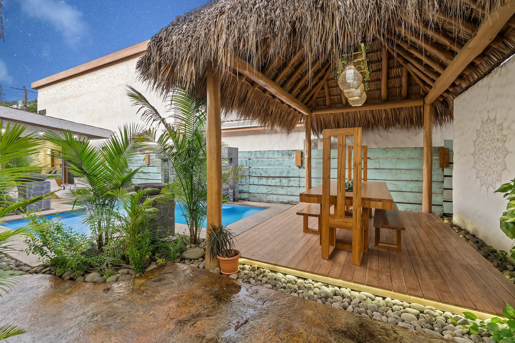 Una zona de comedor abierta con techo de paja da a una piscina y plantas tropicales.