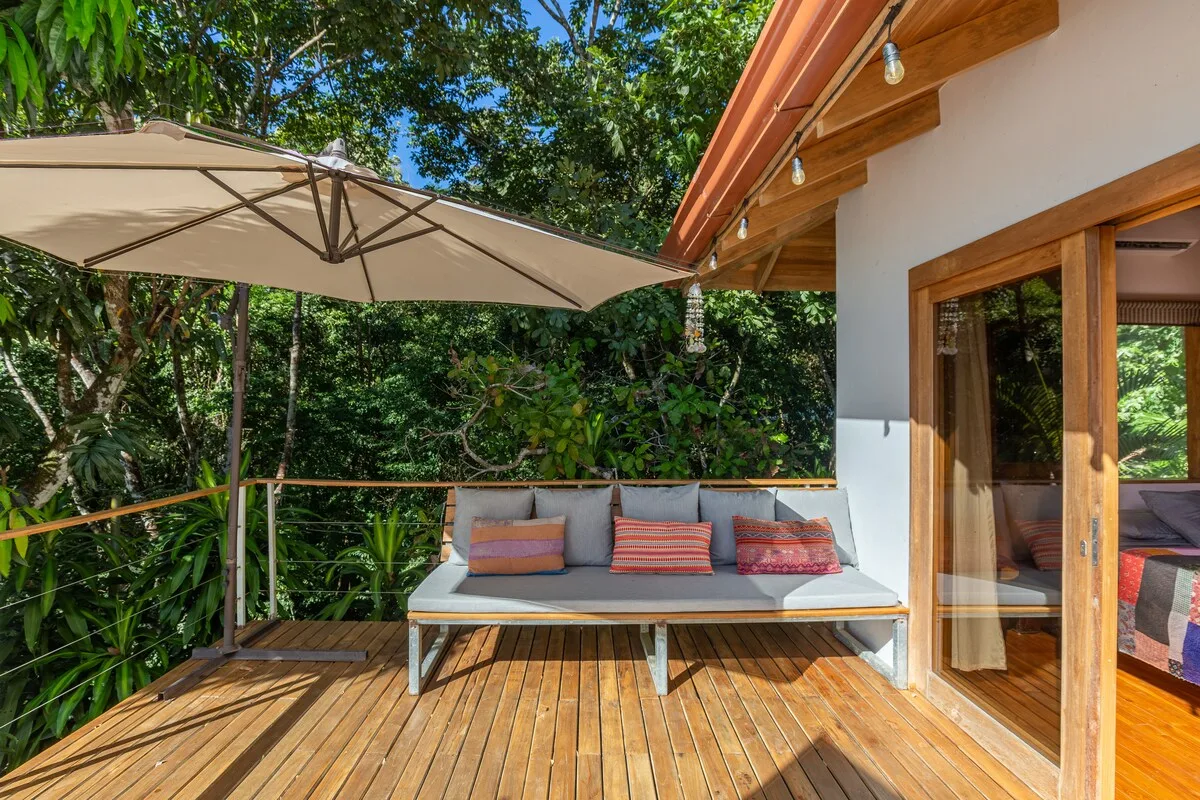 Una terraza exterior con asientos acolchados bajo una sombrilla grande, rodeada de vegetación.
