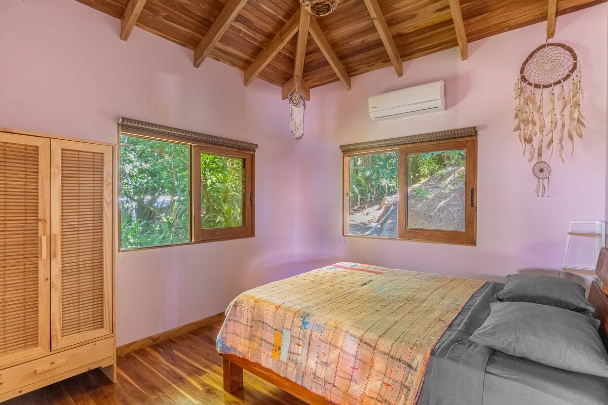 Un dormitorio con una cama bajo un techo de madera y dos ventanas con vistas a la vegetación.