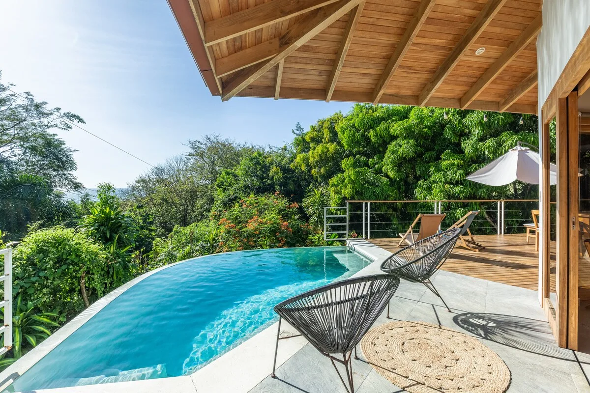 Una piscina curva junto a un patio de madera con asientos y abundante vegetación.