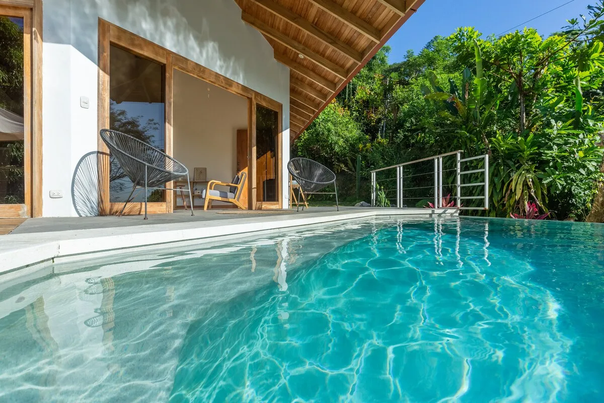 Una piscina junto a un patio cubierto, con vegetación alrededor del área exterior.