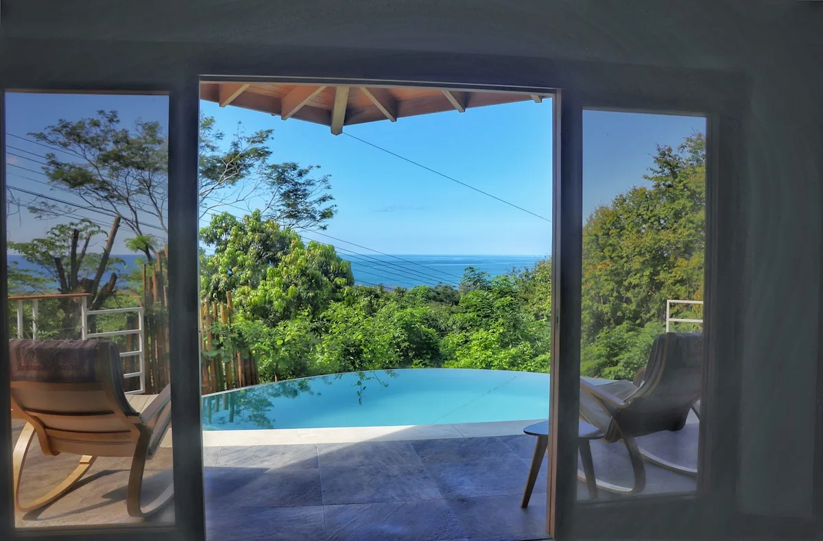 Un patio se abre a una piscina, con árboles y el océano al fondo.