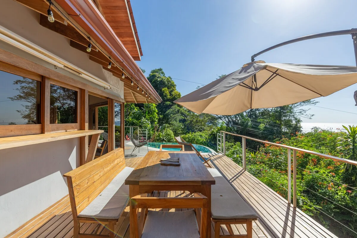 Una terraza con mesa de madera, asientos y una sombrilla grande.