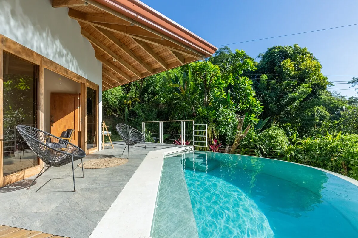 Una piscina turquesa junto a un patio cubierto con asientos y abundante vegetación.
