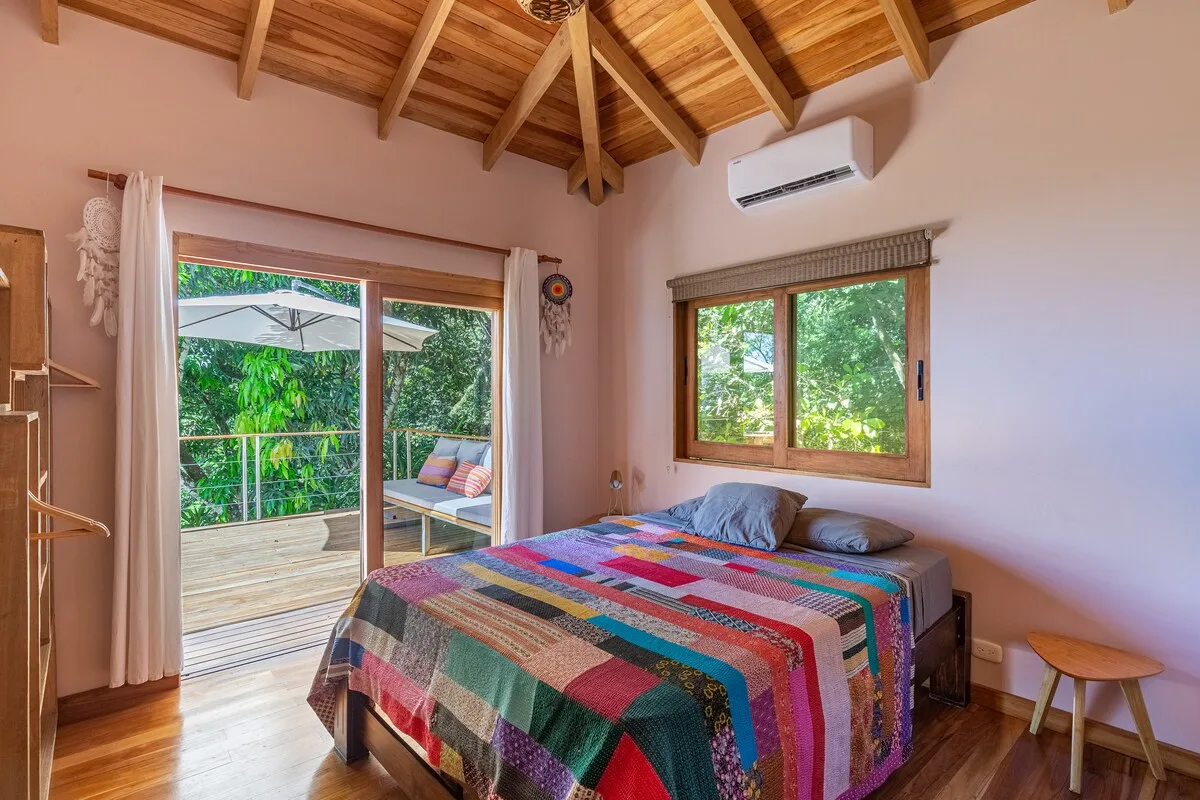 Un dormitorio con una colcha colorida, techo de madera y ventanas con vistas a la vegetación.