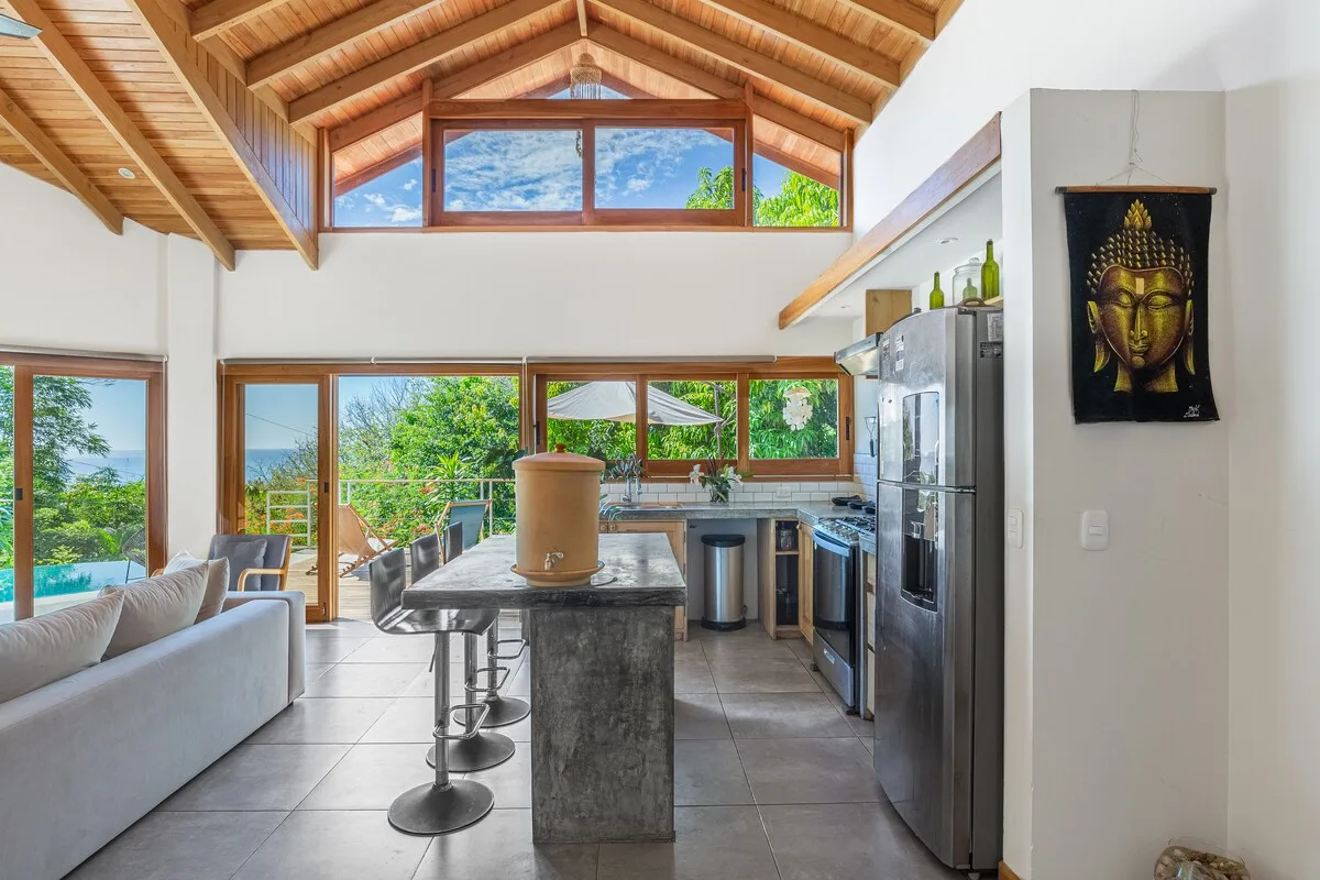 Una luminosa sala de estar y cocina abiertas, con grandes ventanas y techo abovedado de madera.