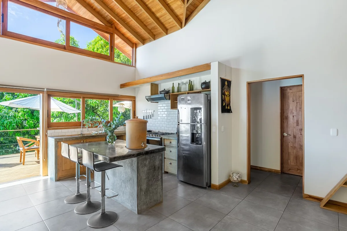 Una cocina abierta y luminosa con una isla central y grandes ventanas con vistas a la vegetación.