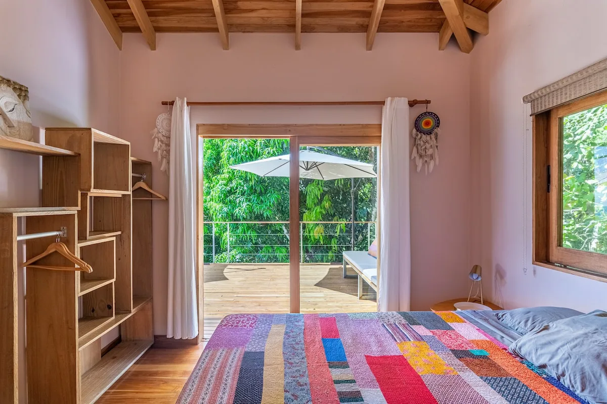 Un dormitorio con una colcha colorida, estantes abiertos y puertas de vidrio hacia una terraza.