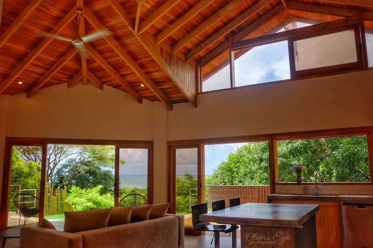 Una sala de estar y comedor con techo alto de madera y ventanas con vistas a la vegetación.
